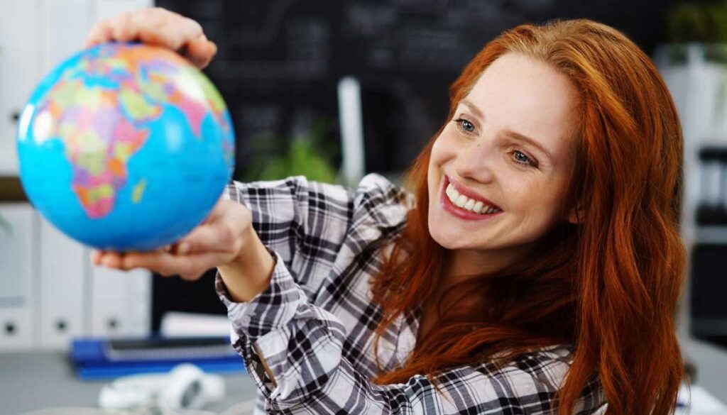 A smiling woman with long red hair in a plaid shirt holds up a small globe, pointing at Africa, with an office setting blurred in the background.