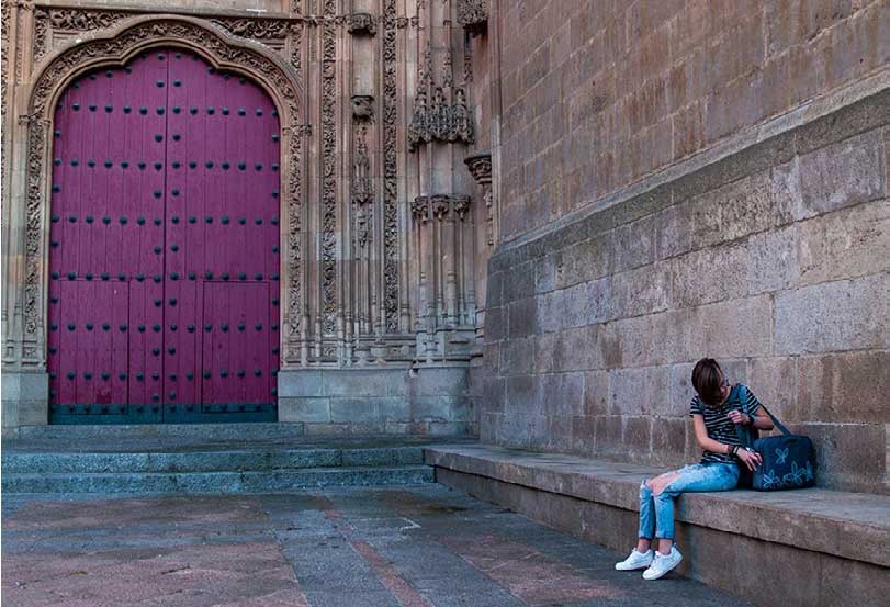 A person with short hair, wearing a striped shirt and ripped jeans, sits on a stone bench near an ornate wall and a large, red, arched door, looking into their bag.
