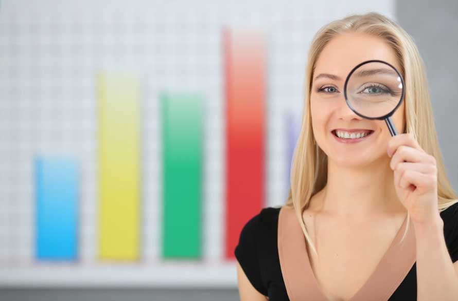 A smiling woman holds a magnifying glass to her eye, standing in front of a blurry bar graph with yellow, green, blue, and red bars on a white grid background.