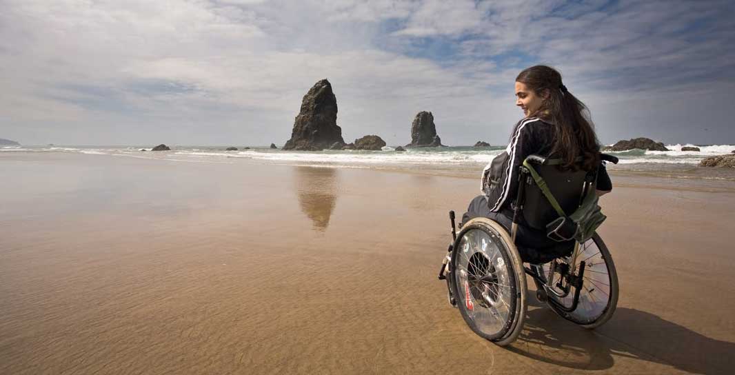 A woman in a wheelchair sits on a sandy beach near the water, looking toward large rock formations in the ocean under a partly cloudy sky.