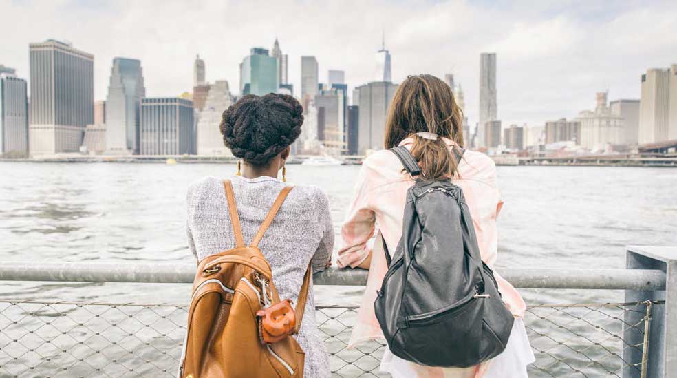 Two people with backpacks stand by a waterfront fence, looking at a city skyline with tall buildings across the river on a cloudy day.