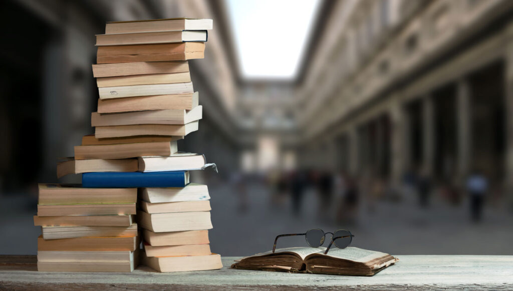 A stack of books sits on a wooden surface next to an open book with eyeglasses on top. In the background, blurred classical architecture and people are visible, giving the scene an academic or scholarly atmosphere.