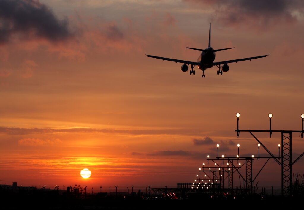 A passenger airplane is approaching a runway for landing at sunset, with the sky glowing orange and runway lights illuminated along the landing path.