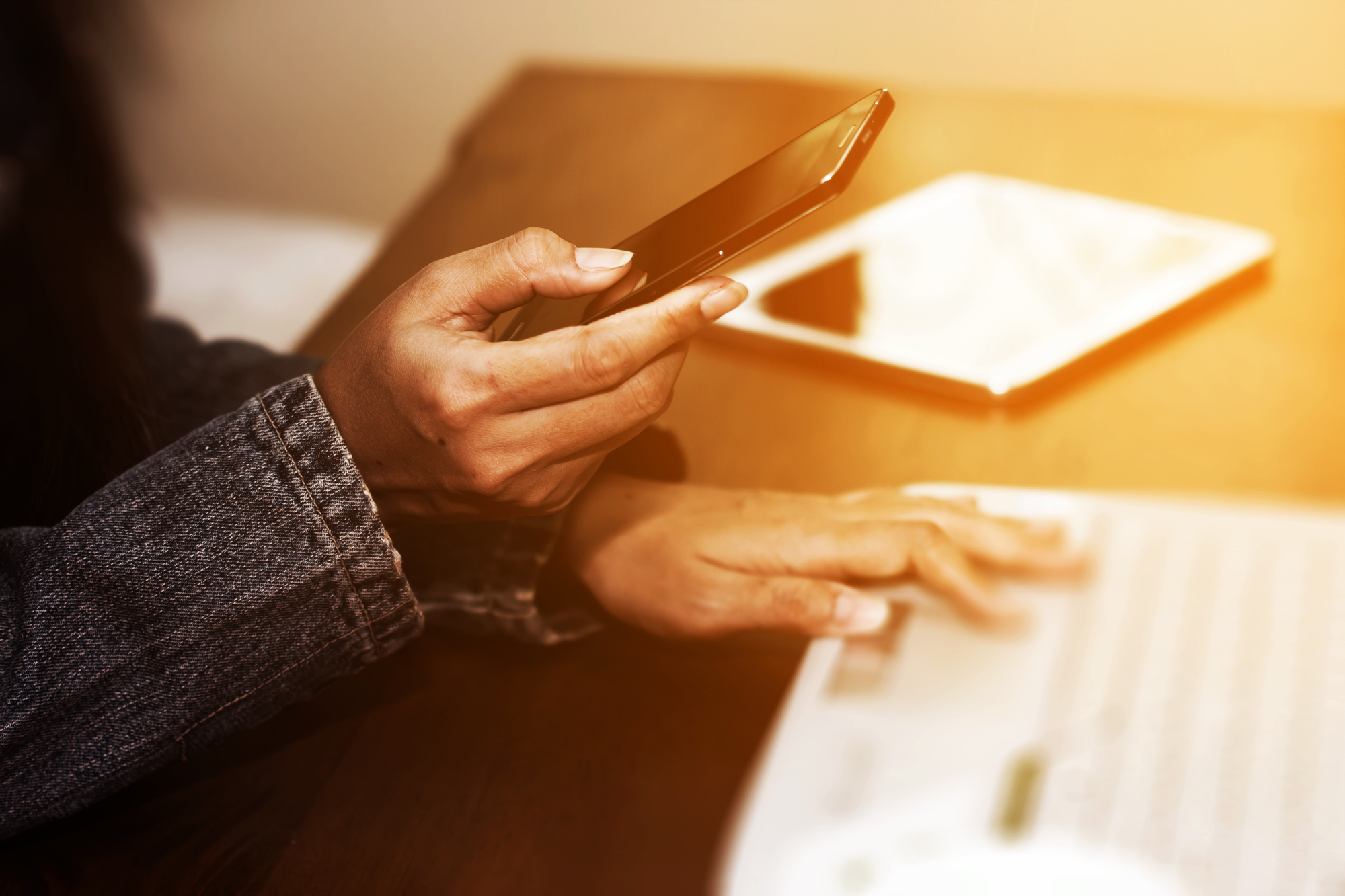 A person wearing a denim jacket holds a smartphone in one hand and types on a laptop keyboard with the other, with a tablet visible on the desk in a warmly lit setting.