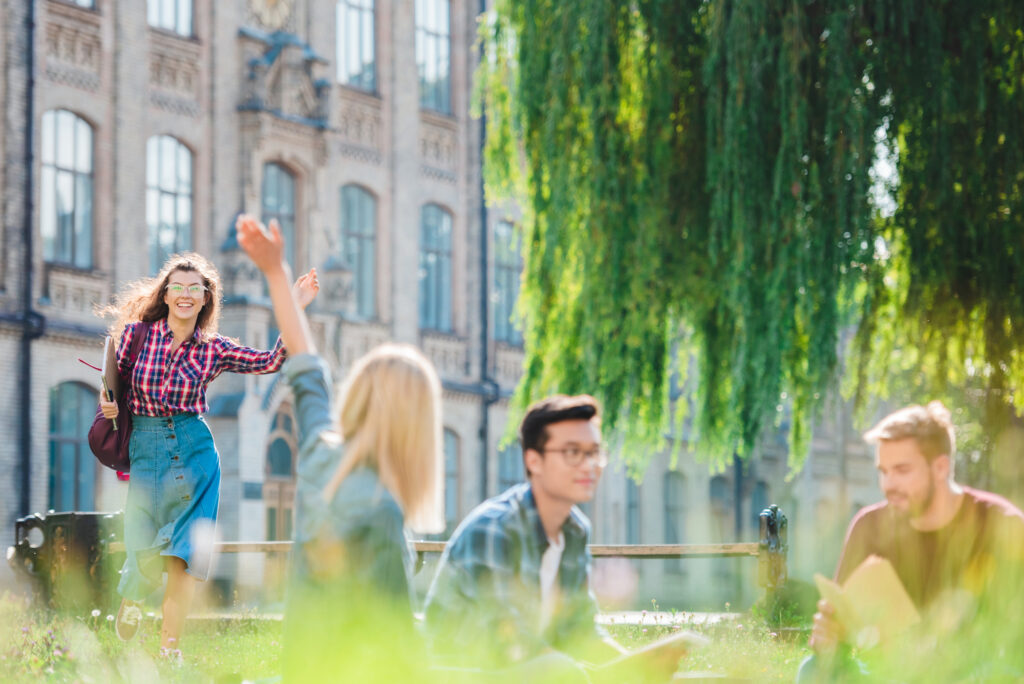 A young woman waves as she approaches a group of three friends sitting on the grass outside a historic building on a sunny day. The setting is bright and relaxed, surrounded by greenery.