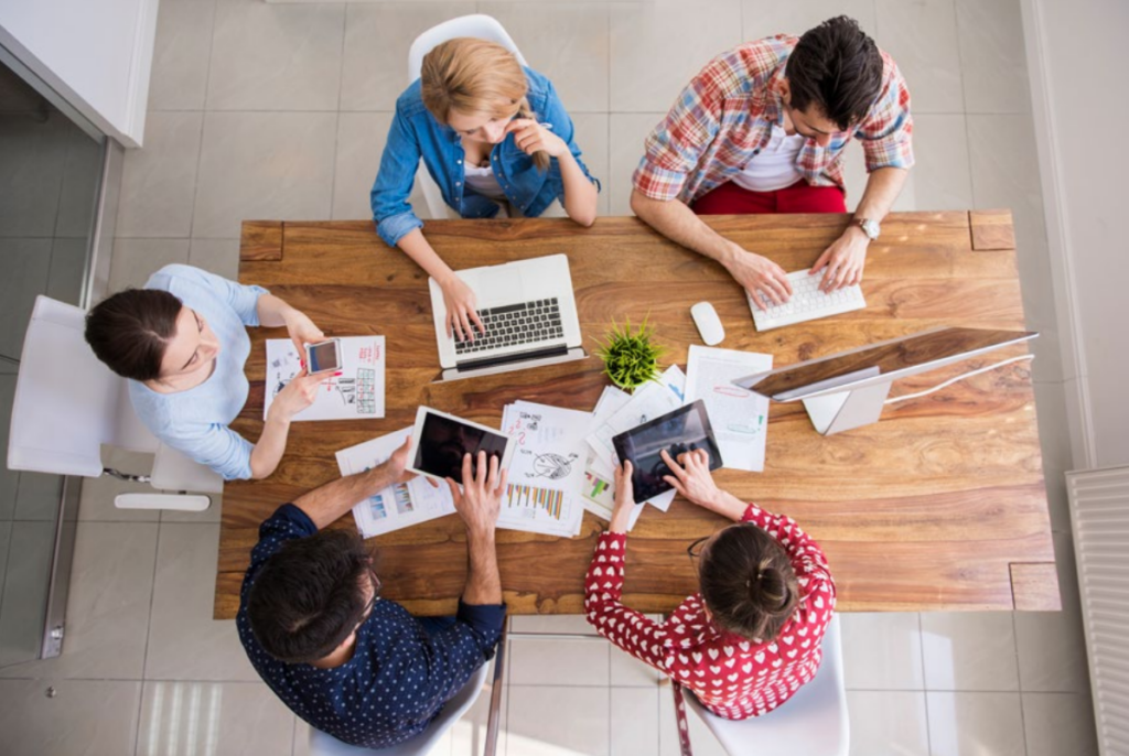 Five people sit around a wooden table working on laptops, tablets, and papers with charts. They appear engaged in a collaborative meeting or brainstorming session in a modern office setting, viewed from above.