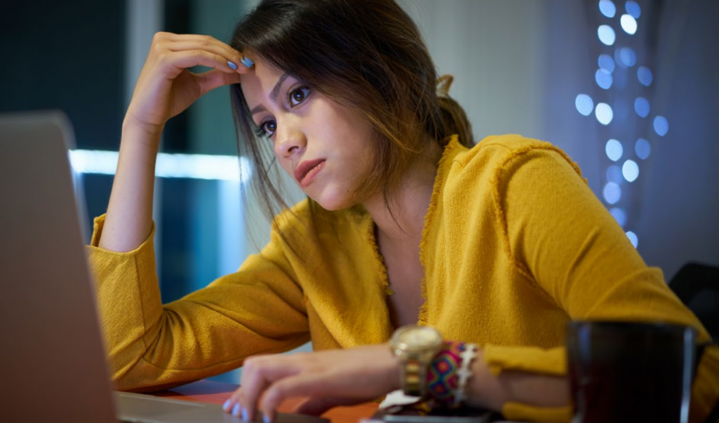 A woman in a yellow top sits at a desk, looking thoughtfully at a laptop screen with her hand resting on her forehead. The background is softly lit with blurred string lights.