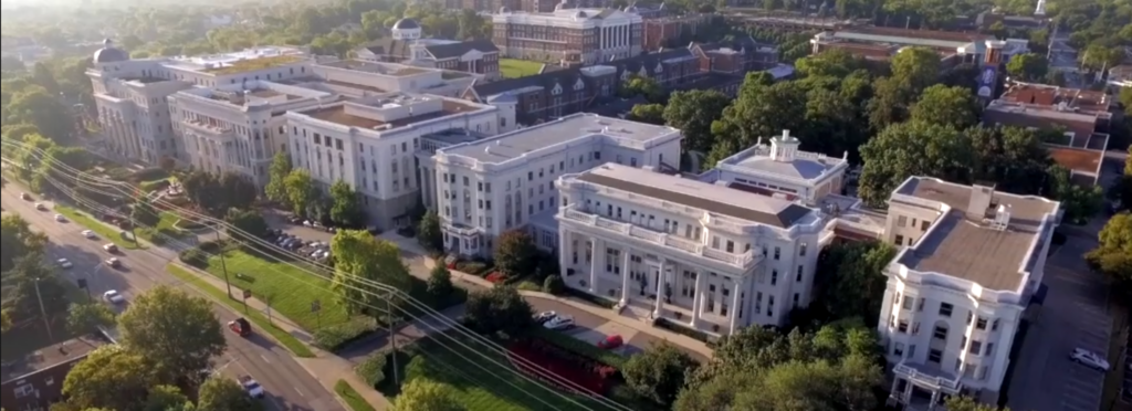 Aerial view of a large university campus with multiple white historic buildings surrounded by trees and green lawns, adjacent to a street with cars and power lines.