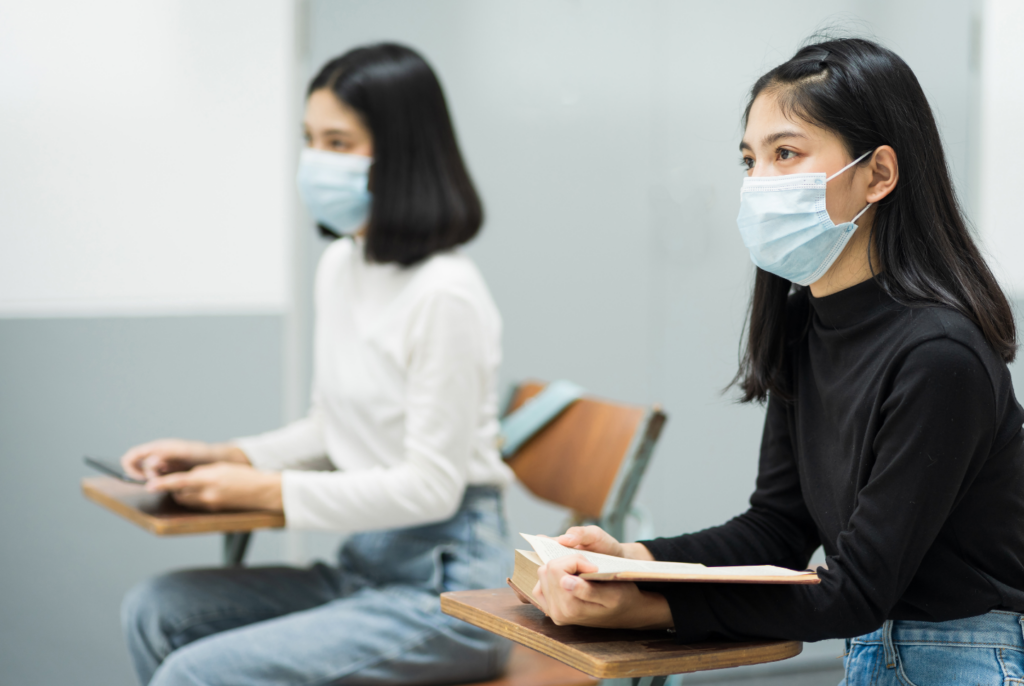 Two students wearing face masks sit at desks in a classroom. One student holds a book, while the other uses a tablet. Both are focused, with some distance between them, suggesting adherence to health guidelines.