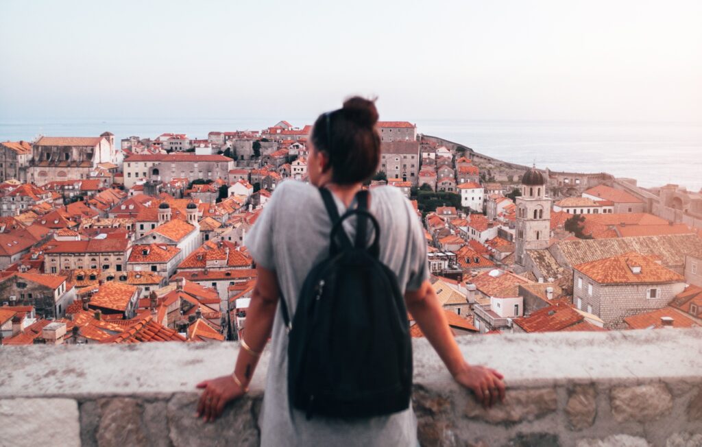 A person with a black backpack stands on a stone terrace, overlooking a scenic cityscape of red-tiled roofs and historic buildings near the sea at sunset.
