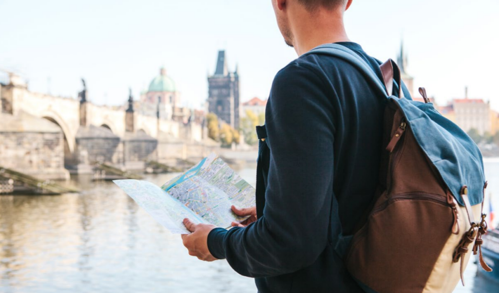 A person wearing a backpack stands by a river, holding a map and looking toward an old bridge and historic buildings in the background on a sunny day.