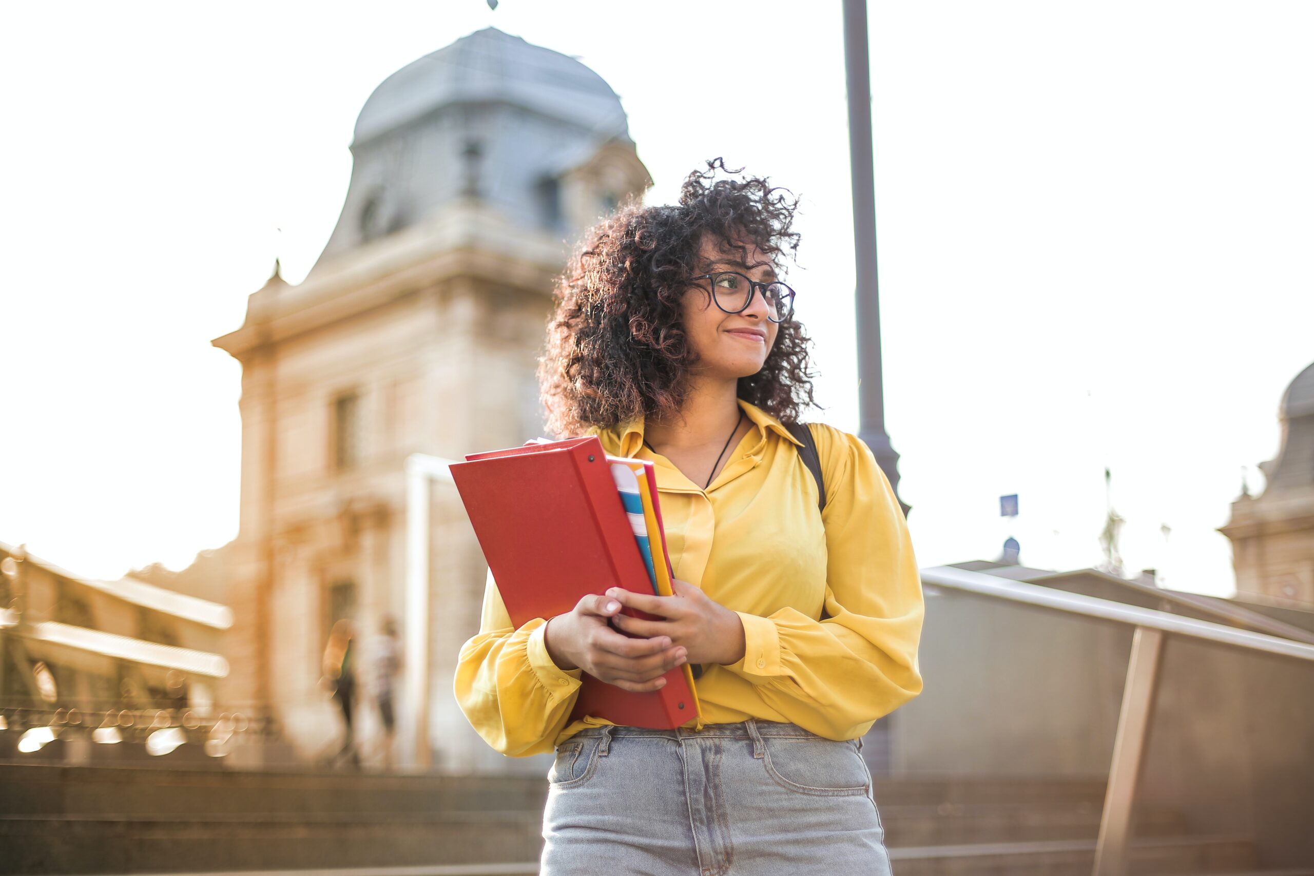 A young woman with curly hair and glasses, wearing a yellow shirt and jeans, holds red folders and books while standing outdoors near historic buildings, smiling and looking to the side.