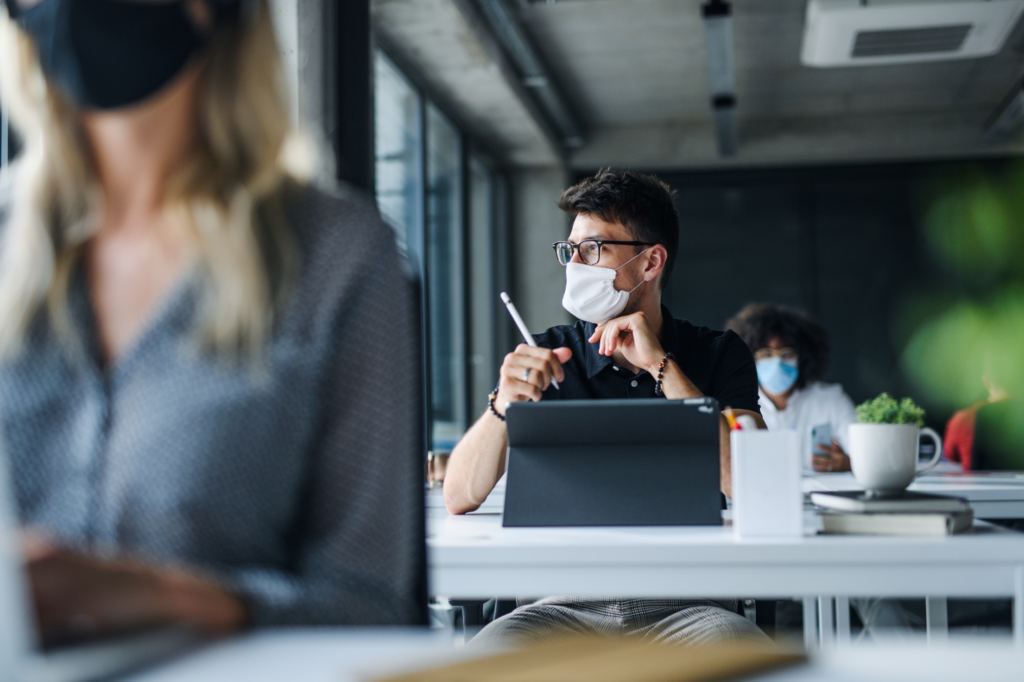 People wearing face masks sit at desks in a modern office. A man in the center, holding a pen, looks to the side. Focus is on him, with blurred foreground and background, suggesting a collaborative work environment.