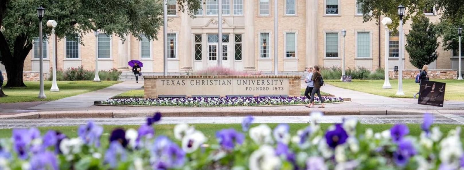 Front view of Texas Christian University’s main building with a stone sign reading Texas Christian University Founded 1873. Purple and white flowers are in the foreground, and two people are walking on the sidewalk.