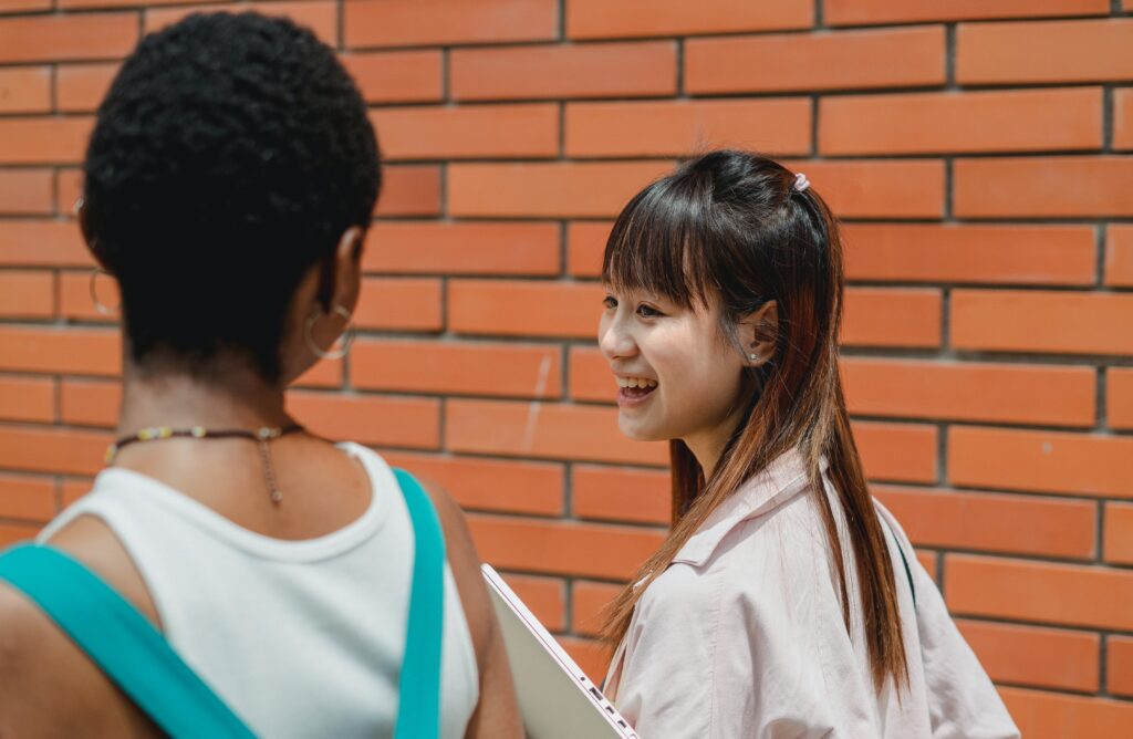 Two young women stand in front of a brick wall, one facing away with short hair and the other smiling, holding notebooks and wearing a light-colored shirt.
