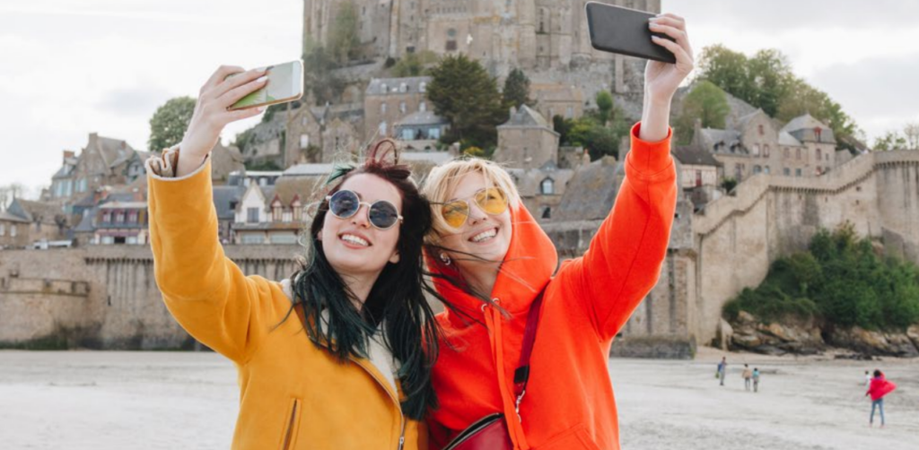Two smiling women in colorful jackets and sunglasses take selfies in front of an old stone castle and village, with other people visible in the background on a cloudy day.