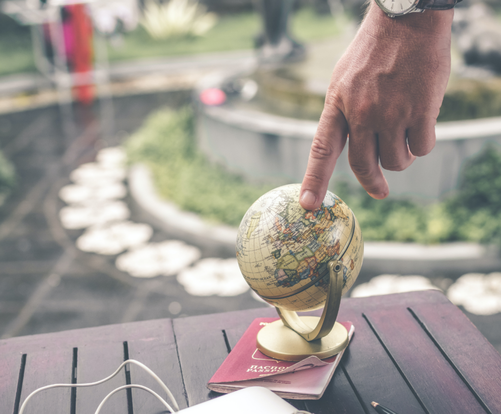 A person points at a small globe on a table next to a passport, with a garden and stone path blurred in the background.