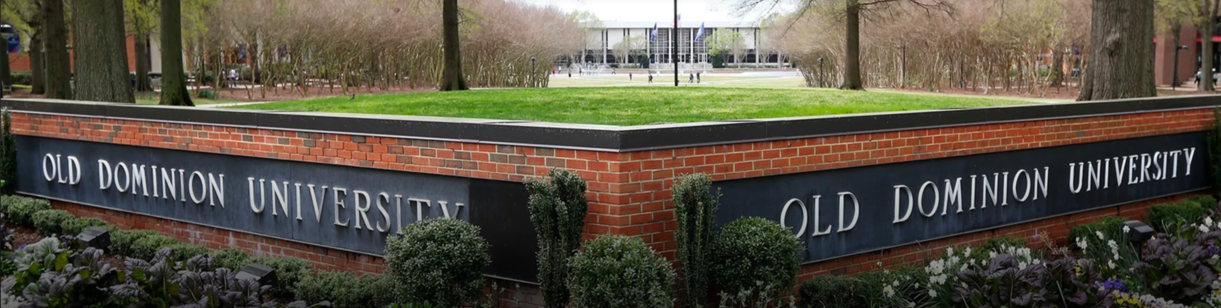 A brick and stone corner sign reads Old Dominion University on both sides, with green bushes in front and a campus building visible in the background amid trees.
