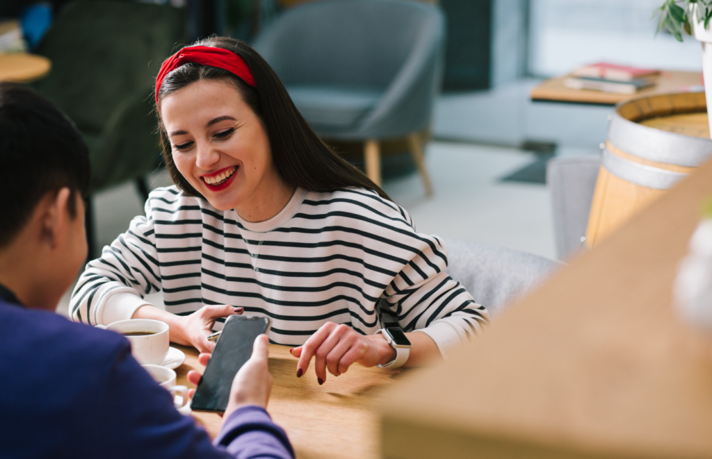 A smiling woman wearing a striped shirt and red headband sits at a cafe table, looking at a phone held by another person, with coffee cups and a cozy atmosphere around them.