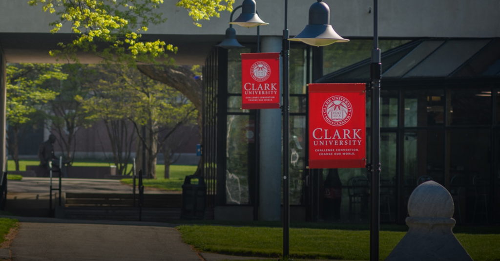 A campus walkway with trees and lamp posts displaying red Clark University banners. A glass building and shaded seating area are visible in the background.