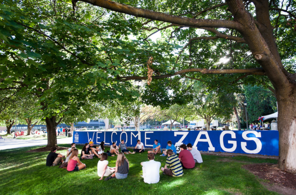 A group of students sits in a circle on green grass under large trees in front of a blue wall painted with “WELCOME ZAGS.” Sunlight filters through the leaves, creating a welcoming, relaxed campus atmosphere.