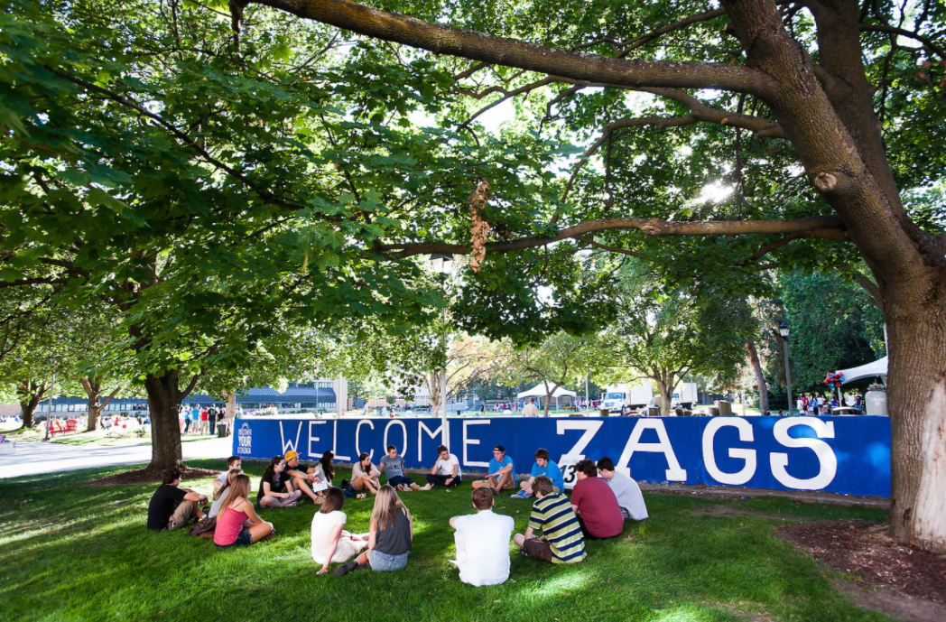 A group of students sits in a circle on green grass under large trees in front of a blue wall painted with “WELCOME ZAGS.” Sunlight filters through the leaves, creating a welcoming, relaxed campus atmosphere.