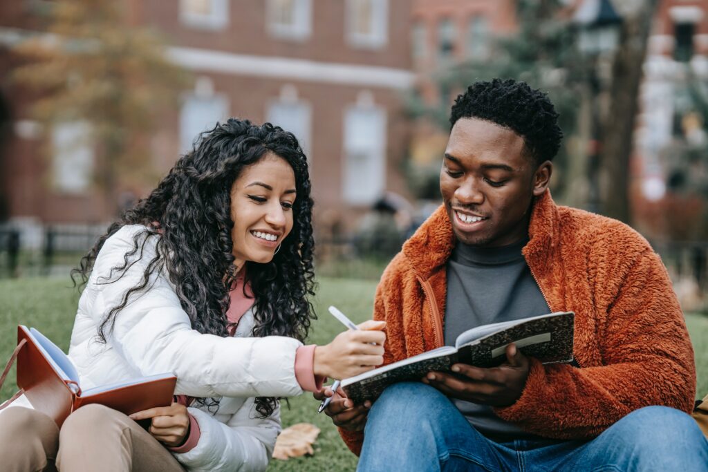 Two smiling students sit on grass outside, studying together. One holds a notebook while the other writes in it, and both appear engaged and happy. Buildings and trees are blurred in the background.