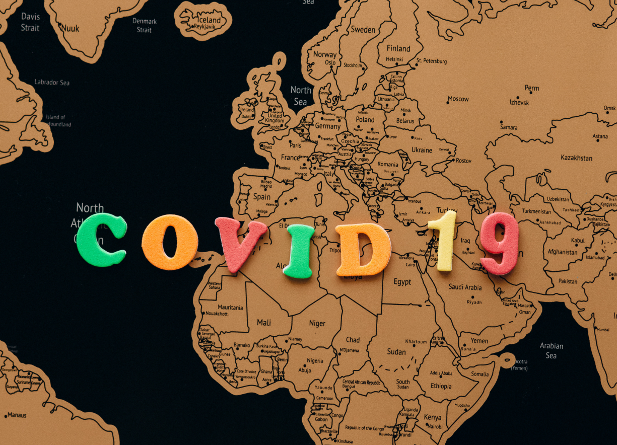 Colorful foam letters spelling COVID 19 are placed on a brown and black world map, with Europe and northern Africa prominently visible in the background.