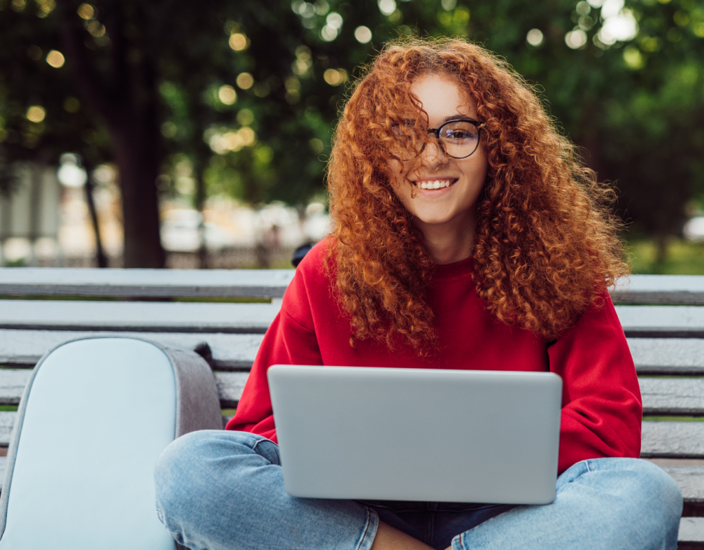 A young person with curly red hair and glasses, wearing a red sweatshirt, sits cross-legged on a bench outdoors with a laptop on their lap and a light blue backpack beside them, smiling at the camera.