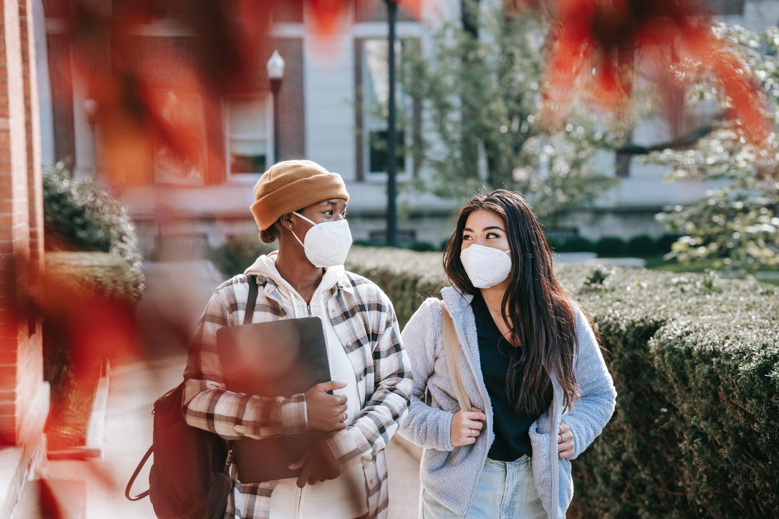 Two students wearing face masks walk outdoors on a campus path, carrying backpacks and a laptop, with autumn leaves and buildings in the background. They are engaged in conversation.
