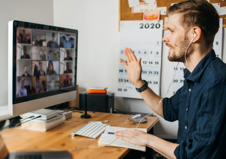 A man wearing earphones sits at a desk, waving at a computer screen displaying a video call with many participants. A 2020 calendar and bulletin board are visible in the background.