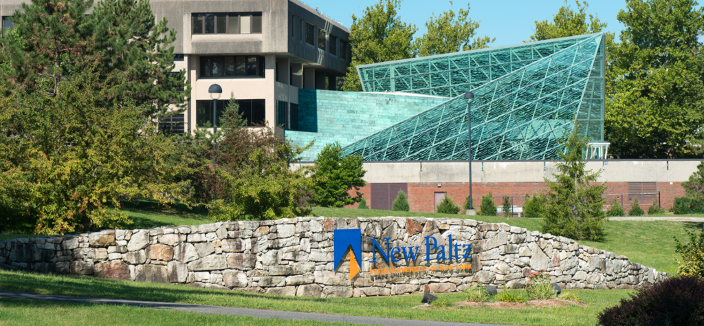A stone sign reading New Paltz with a modern glass and concrete building and lush green trees in the background on a sunny day.