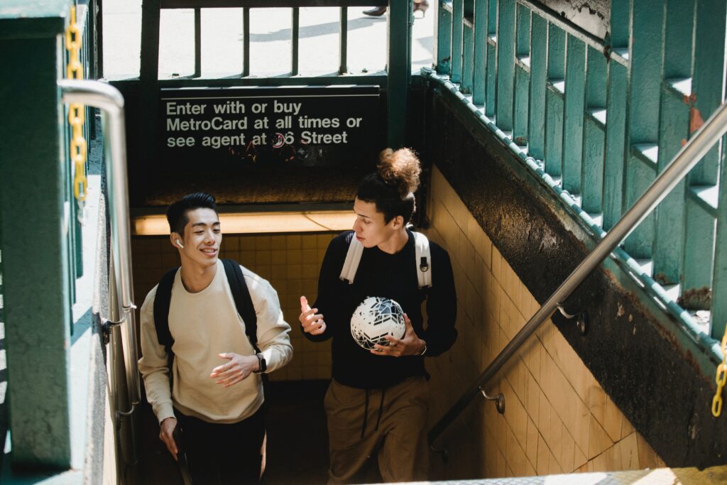 Two young men with backpacks walk up subway stairs. One holds a soccer ball, and both are talking and smiling. Green railings and a MetroCard sign are visible above the stairs. Sunlight shines into the entrance.