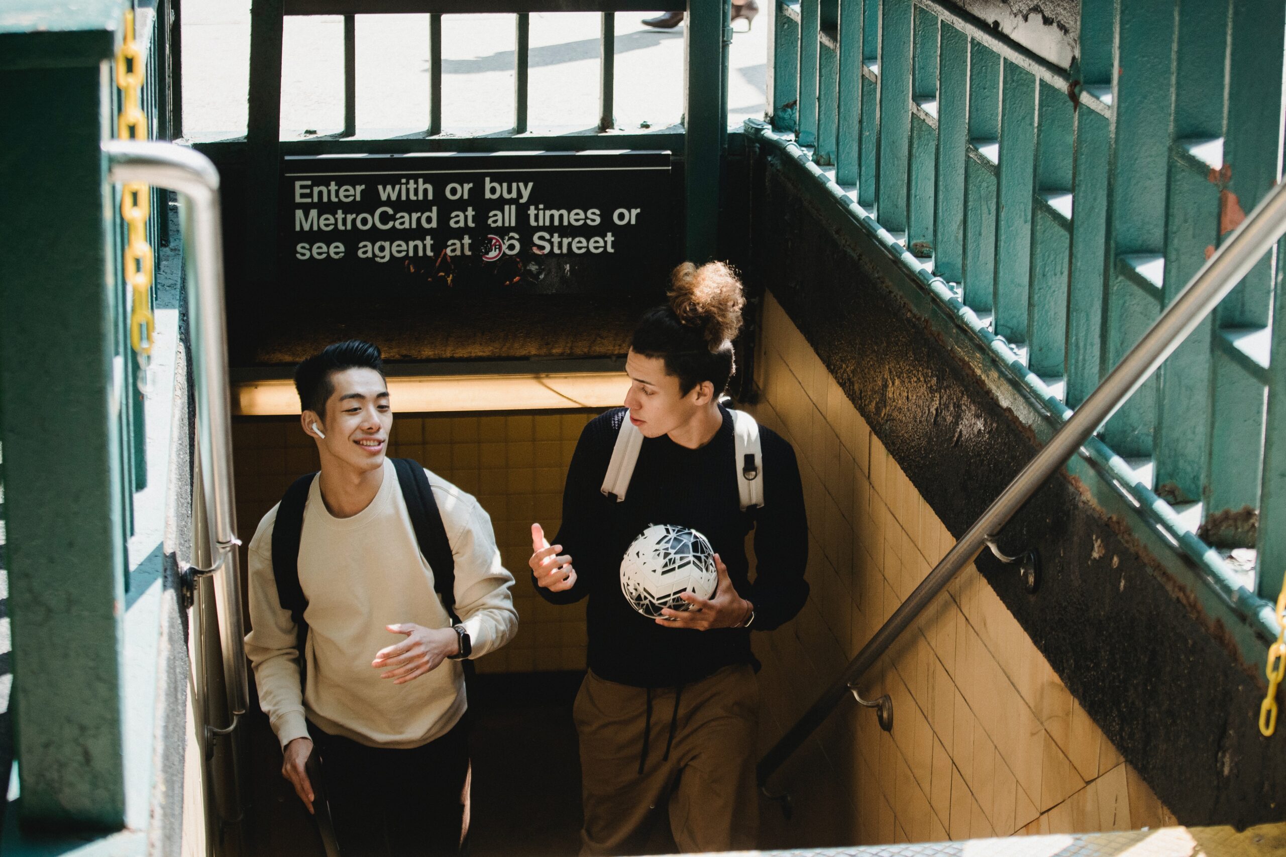 Two young men with backpacks walk up subway stairs. One holds a soccer ball, and both are talking and smiling. Green railings and a MetroCard sign are visible above the stairs. Sunlight shines into the entrance.