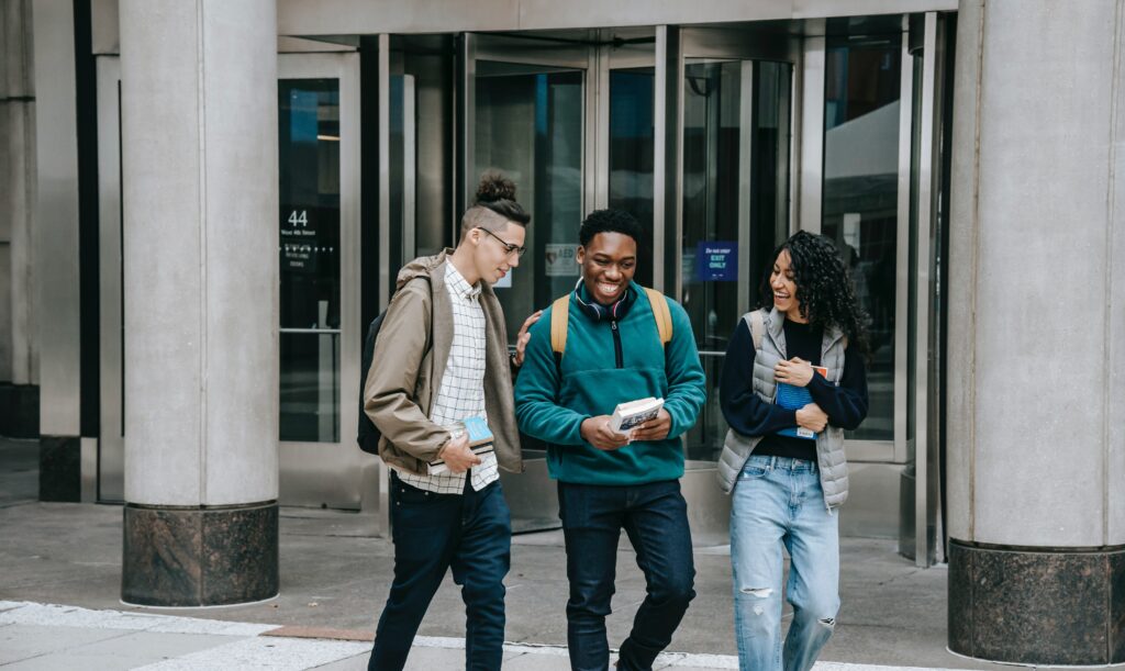 Three students walk and talk outside a building entrance, smiling and holding books and backpacks. They appear to be enjoying each others company on a university or college campus.