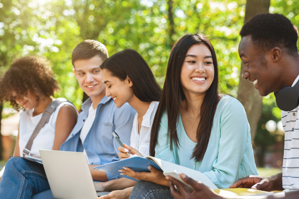 A group of five young adults sit outdoors on a bench, studying and smiling. They have notebooks, a laptop, and headphones, and are surrounded by greenery and sunlight.