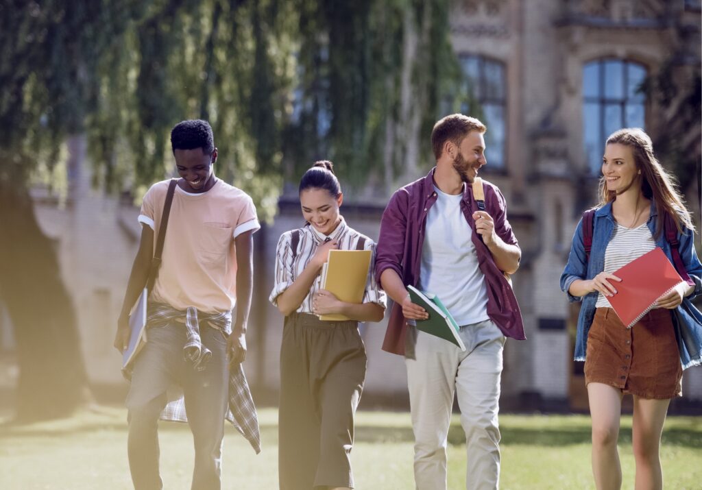 Four college students walk together outdoors, smiling and talking. They carry notebooks and backpacks, with a university building and trees in the background on a sunny day.