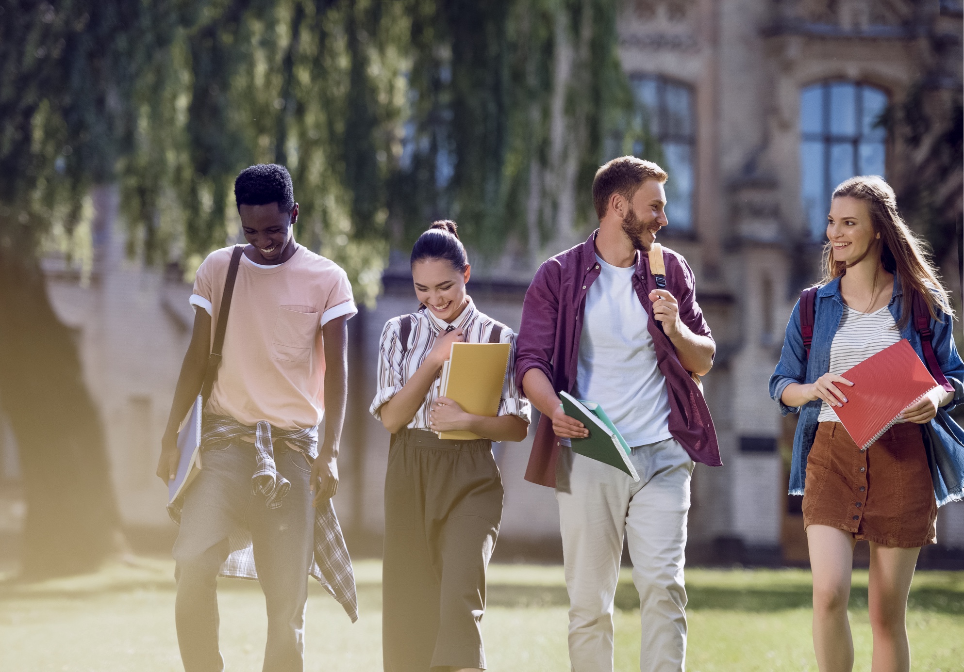 Four college students walk together outdoors, smiling and talking. They carry notebooks and backpacks, with a university building and trees in the background on a sunny day.