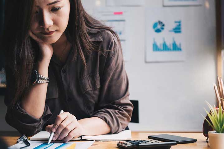 A woman sits at a desk, looking thoughtfully at papers with charts and graphs. She holds a pen in one hand, with a calculator and notebook nearby, and charts are visible on the wall behind her.