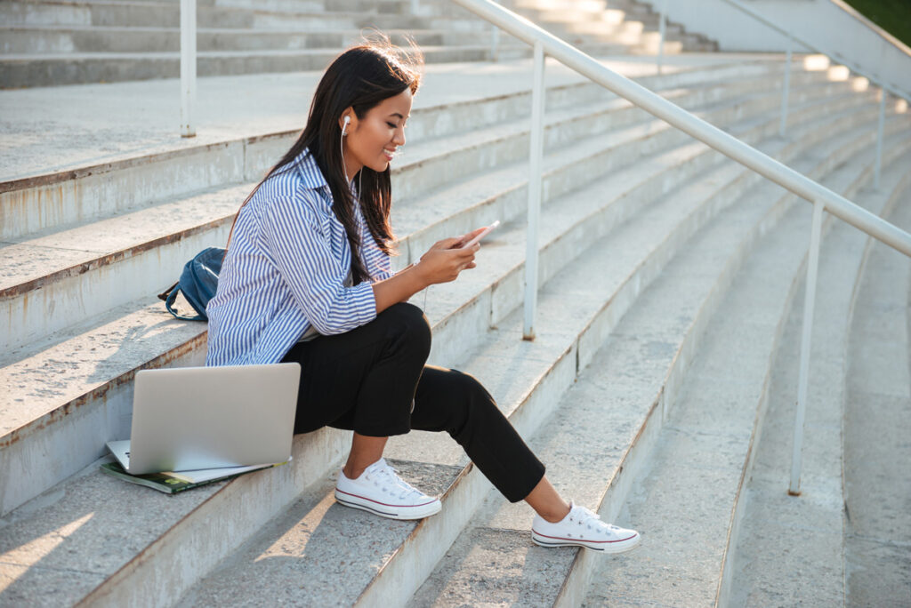 A young woman sits on outdoor steps, wearing earphones and smiling at her phone. She has a laptop, notebook, and backpack beside her, and appears relaxed, enjoying a sunny day.