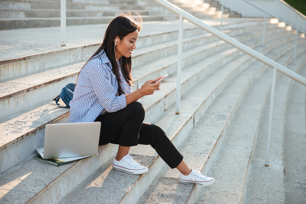 A young woman sits on outdoor steps, wearing earphones and smiling at her phone. She has a laptop, notebook, and backpack beside her, and appears relaxed, enjoying a sunny day.