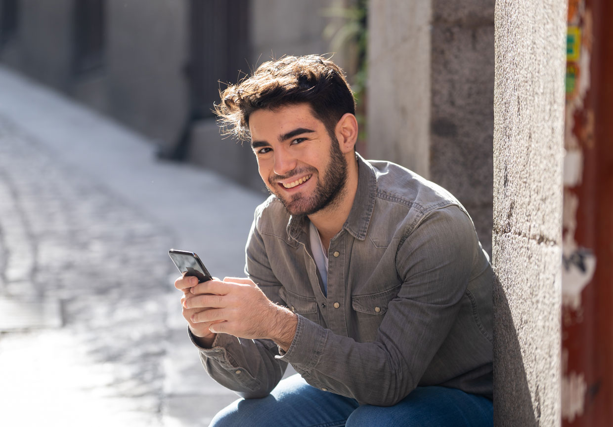 A young man with a beard, wearing a gray shirt and jeans, sits outside on a sunny day, smiling and holding a smartphone in his hands.
