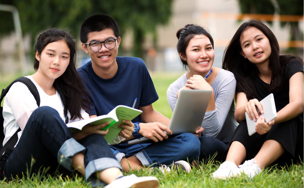 Four young adults sit together on grass outdoors, smiling at the camera. They have books, a laptop, and a notebook, suggesting they are studying or working together in a relaxed, natural setting.