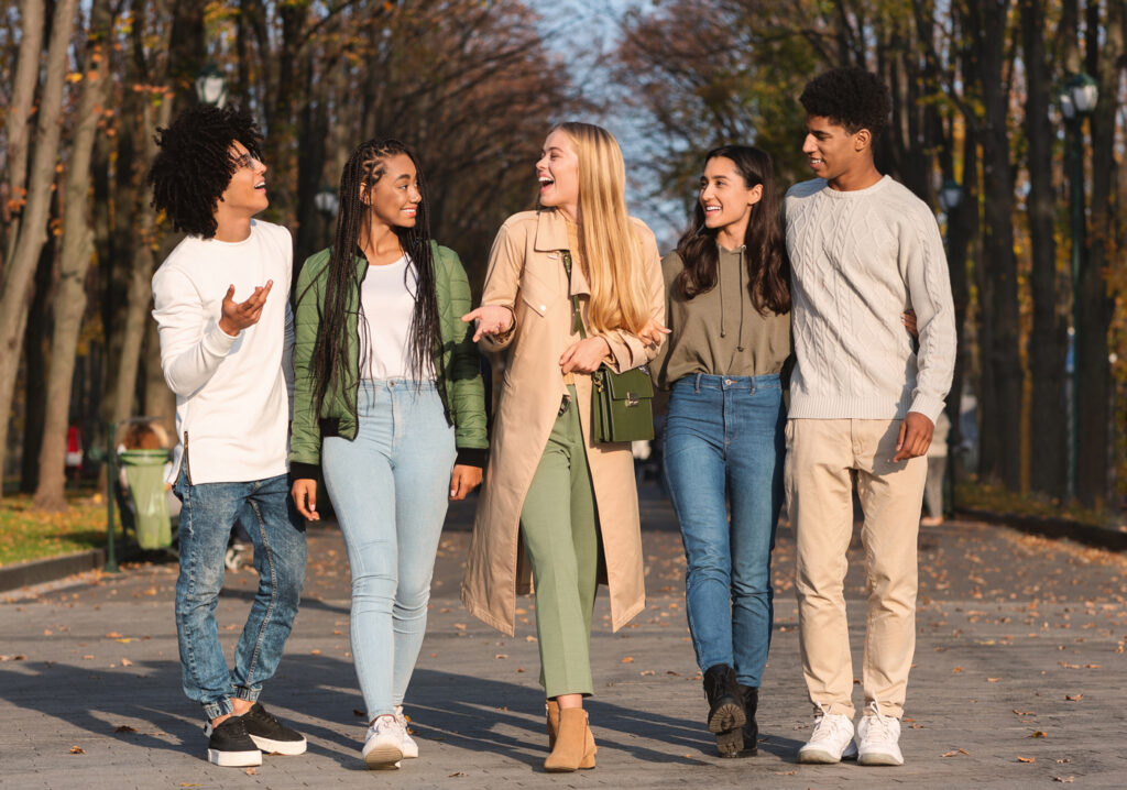Five young adults walk together outdoors on a tree-lined path, smiling and talking. They are dressed in casual fall clothing and appear to be enjoying each others company on a pleasant day.