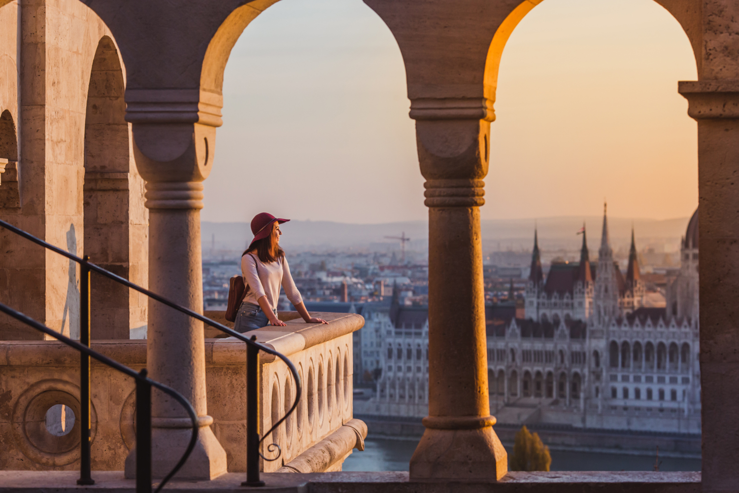 A woman in a hat stands on a stone balcony at sunset, overlooking the city of Budapest with the Hungarian Parliament Building visible in the background through arched columns.