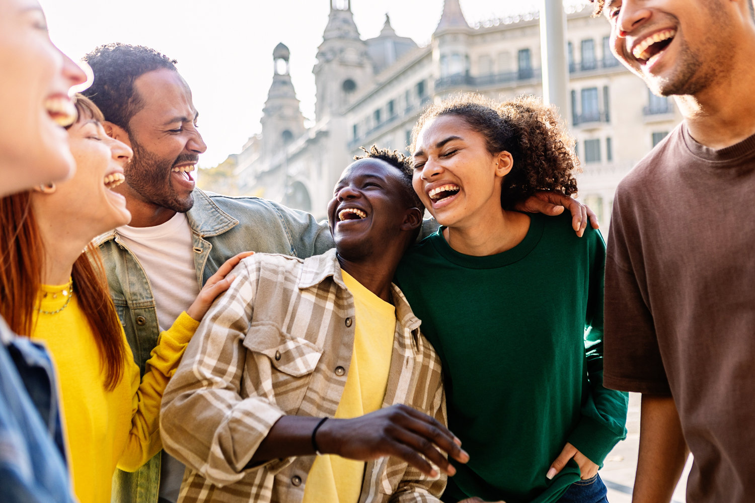 A diverse group of young adults standing close together outdoors, smiling and laughing with arms around each other, with historic buildings in the background on a sunny day.