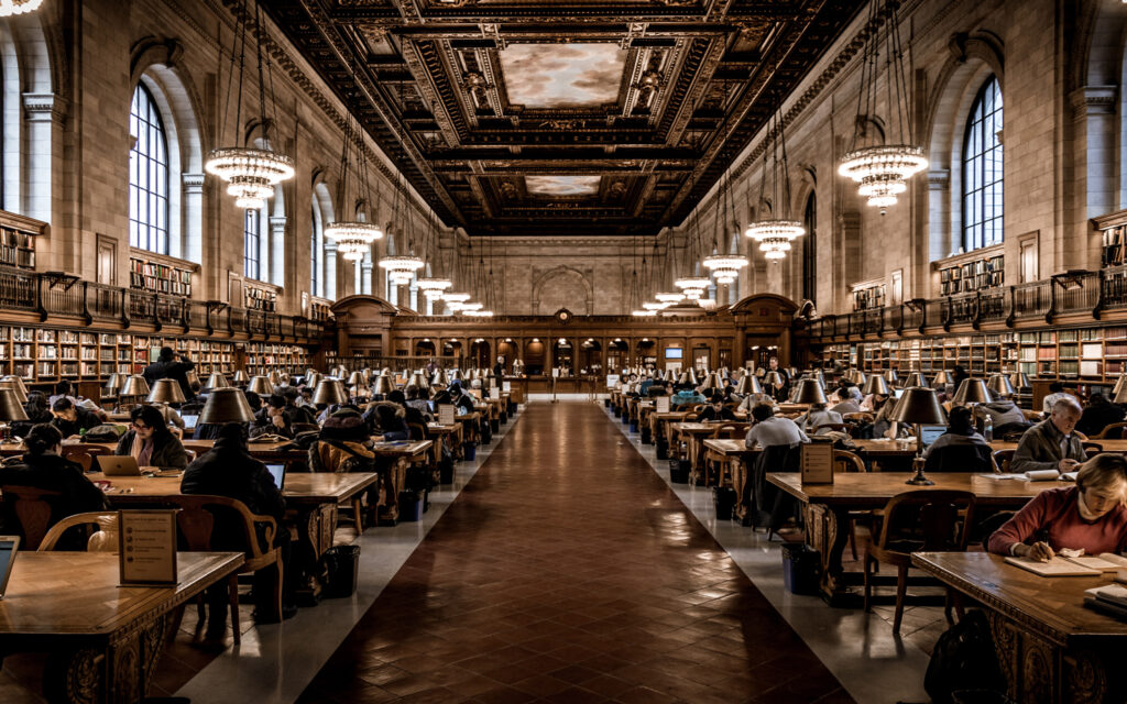 Grand reading room in a large, ornate library with chandeliers, high ceilings, and rows of wooden tables where people are studying and reading under green-shaded lamps. Shelves of books line the walls.
