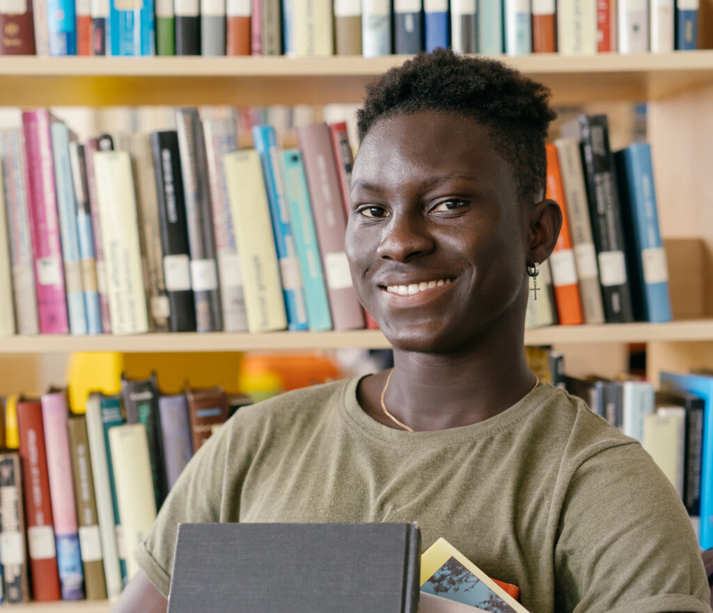 A person smiling and holding books stands in front of a bookshelf filled with colorful books in a library.