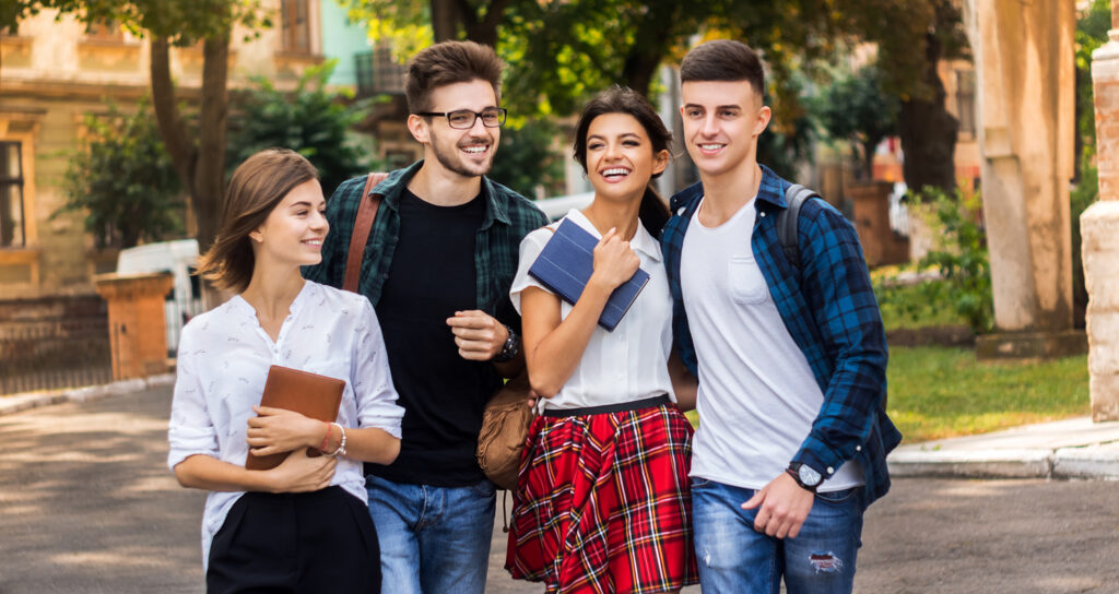 Four young adults walk outdoors, smiling and talking. Two hold books and notebooks, and all are dressed casually. Trees, sunlight, and buildings are visible in the background, giving a campus-like atmosphere.