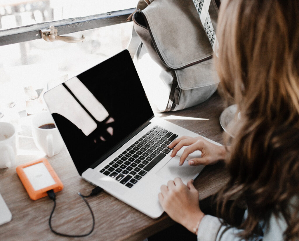A person with long hair works on a laptop at a wooden table next to a backpack, coffee mugs, and an external hard drive connected to the computer.